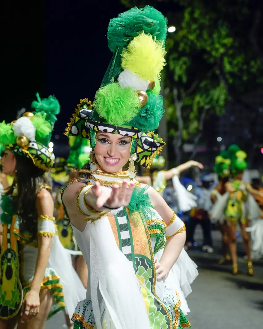 Naty en un desfile de Carnaval Natalia Ferrer con traje de Carnaval verde y blanco y un gran tocado de plumas, extendiendo la mano hacia la cámara durante un desfile nocturno