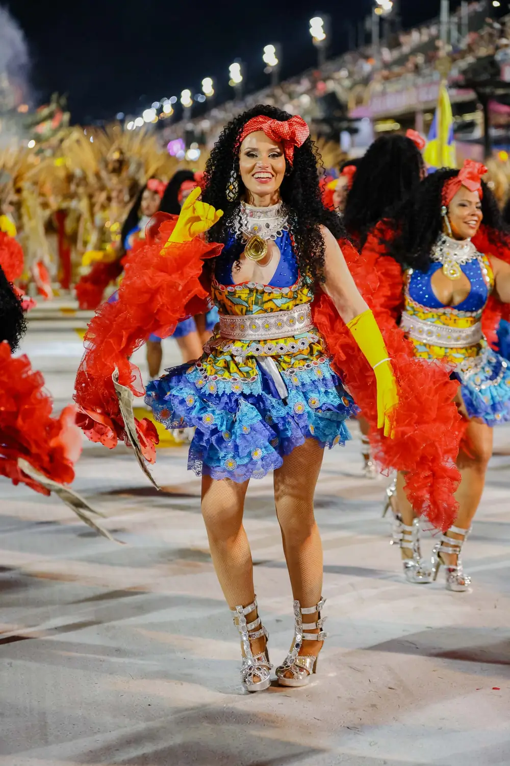 Naty desfilando por la avenida del Sambódromo con traje de Carnaval azul y rojo Naty desfilando de noche en un desfile de Carnaval, con traje azul y amarillo y mangas de tul rojo, entre otras bailarinas con el mismo traje