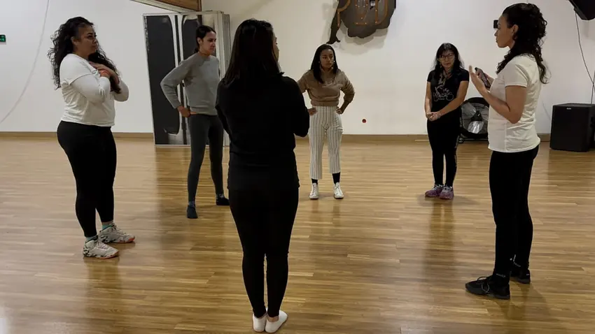 Naty marcando un paso frente a un grupo de mujeres en una sala de baile Naty explicando a un grupo de mujeres de pie en ronda, en un salón de piso de madera