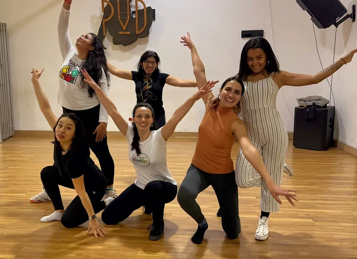 Un grupo de mujeres posando sonrientes con los brazos abiertos en un salón de piso de madera