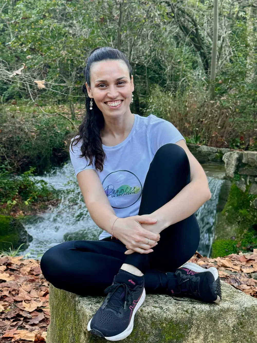 Naty sentada sobre una piedra junto a un arroyo en el bosque, en ropa deportiva, sonriendo